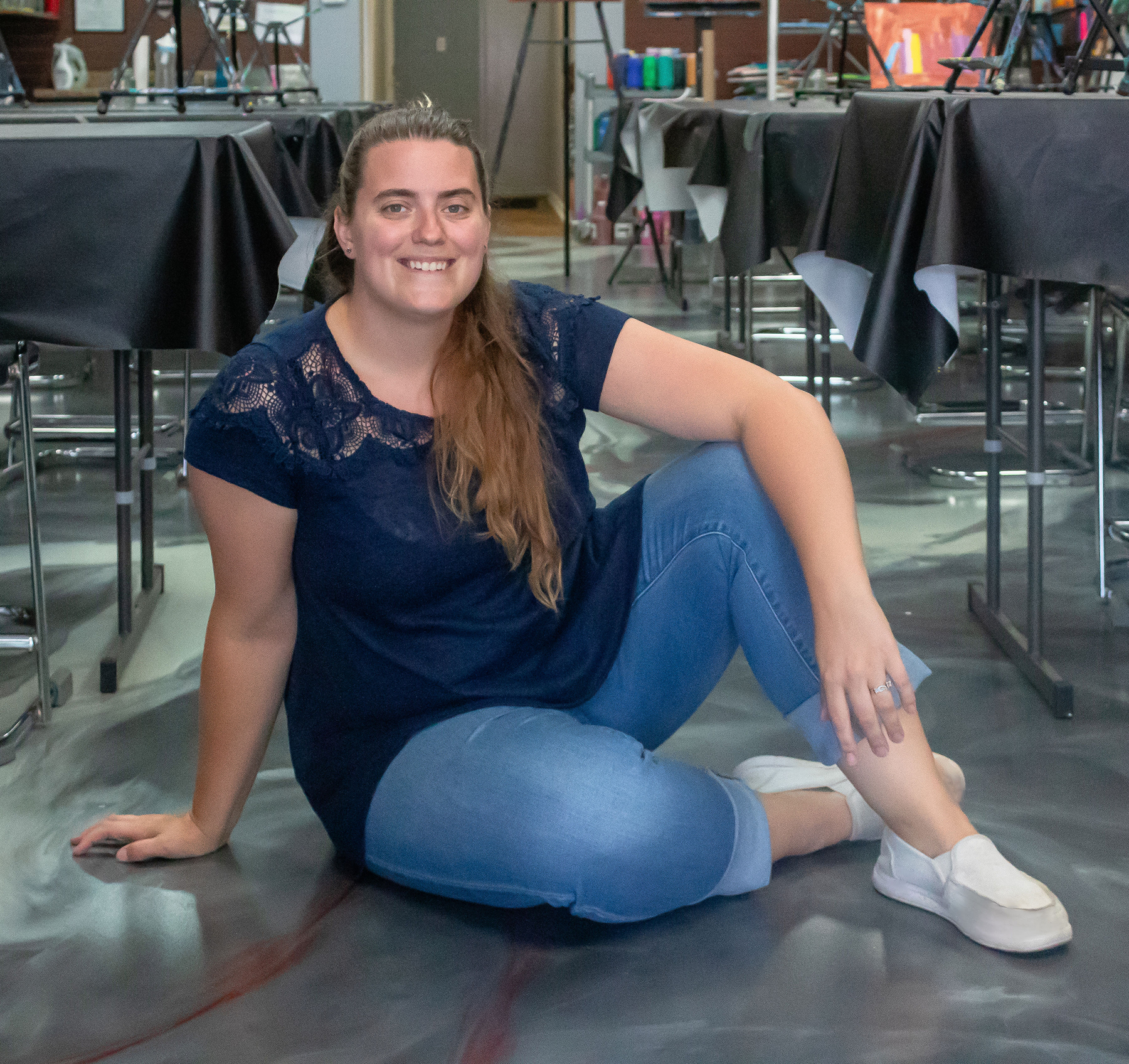 Lauren-Salza-Cheers-N-Paint-Art-Teacher-In-Cary-Nc Lauren Salza, Cheers N Paint Studio Owner And Teacher In Cary, Nc Sitting On The Floor After An Art Class Wearing A Blue Shirt.